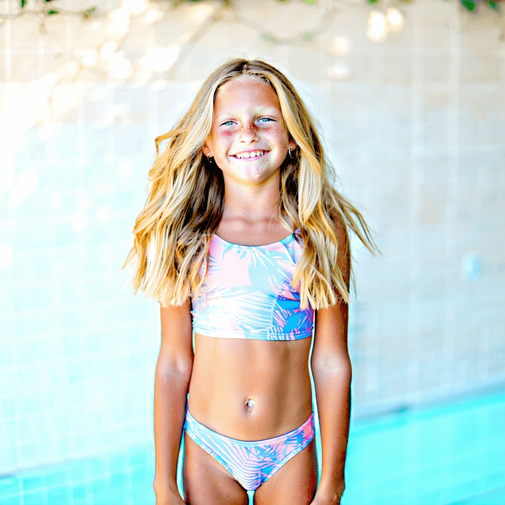 Create a highly detailed, 8k resolution photographic image capturing a full-body view of a cute teen girl with blonde hair. She is standing in a playful pose, embodying a moment of carefree youth, wearing a two-piece swimwear in pastel tones. The scene is bathed in soft natural lighting that highlights her genuine smile and the dynamic motion of her hair, suggesting a gentle breeze. The composition adheres to the rule of thirds, with a shallow depth of field focusing on her, creating a cinematic feel. The perspective is a low angle shot, adding a sense of grandeur and focus on her joyful expression. The environment surrounding her should complement the mood without distracting, ensuring the textures and colors feel realistic and natural.